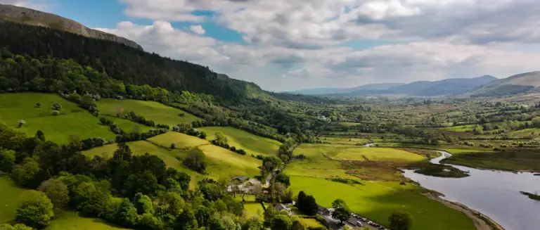 Expansive Irish farm landscape under a cloudy sky, showing fields partitioned by hedges and stone walls, sloping down a hillside toward a river or lake.